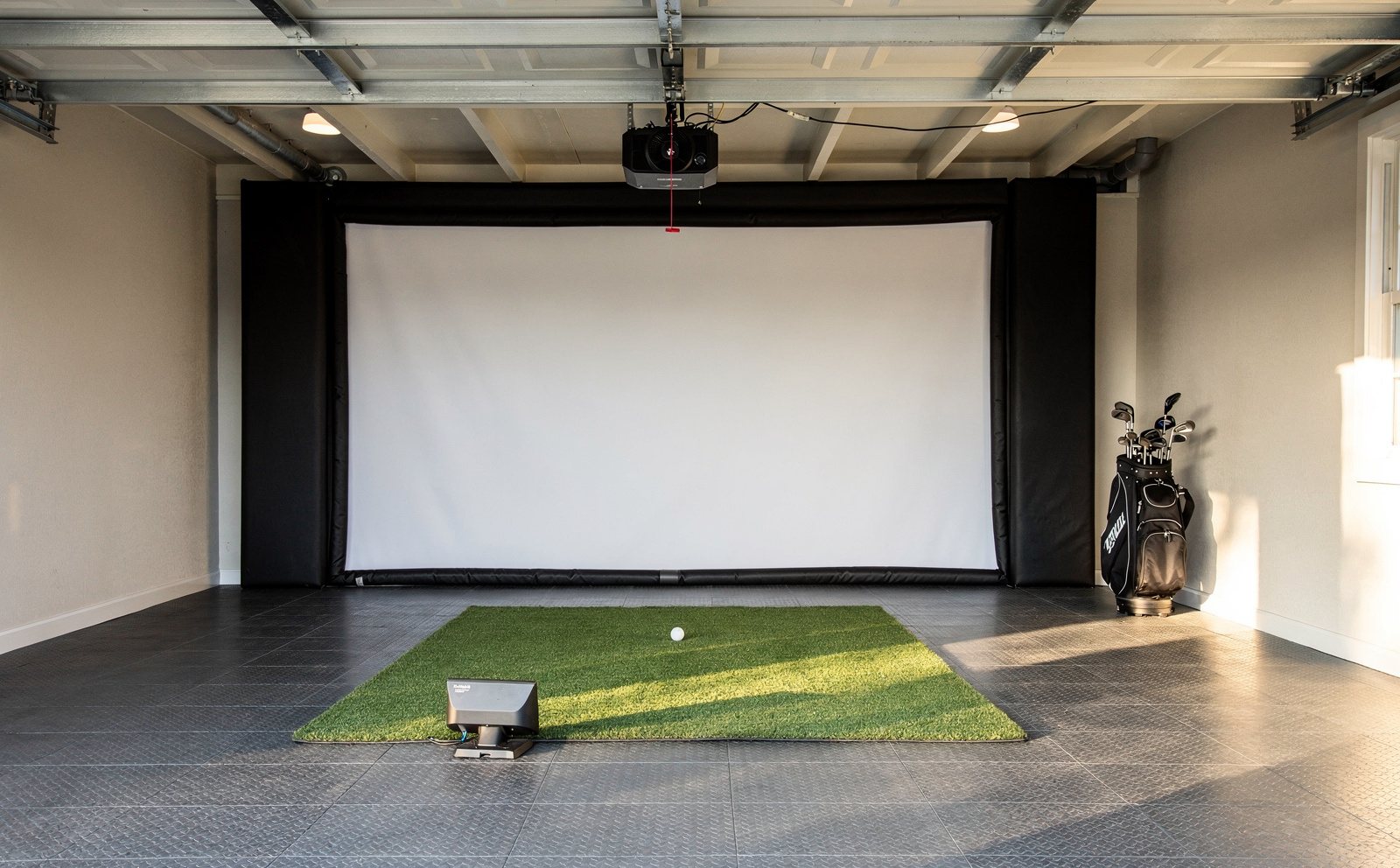 A finished golf simulator bay built into a home garage: impact screen and enclosure at the far wall, hitting mat on tiled flooring, projector mounted overhead