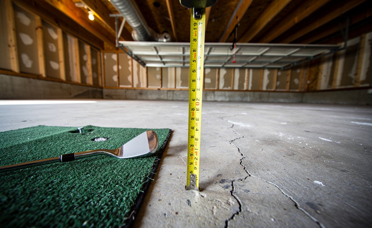 A tape measure held up to the ceiling joists of an unfinished garage, with a golf club and a hitting mat on the floor below